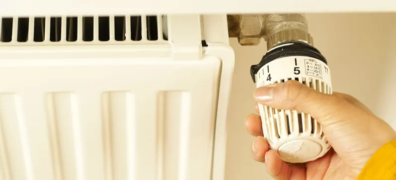 Une main tourne la tête thermostatique d'un radiateur blanc, graduée de 1 à 5